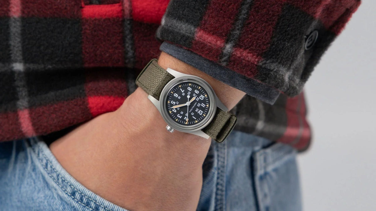 Field watch with an olive fabric strap worn on the wrist.
