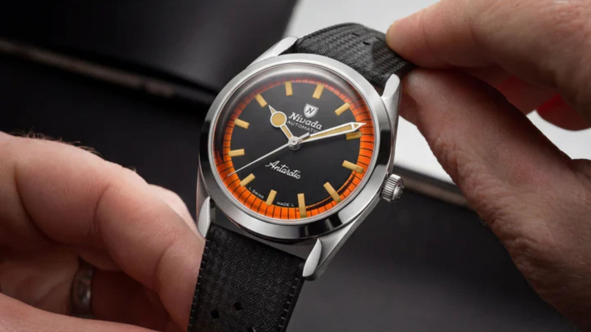 A watch being held by two hands, showing a steel case, a matte black dial with a vibrant orange checkered outer ring, and a black textured rubber strap