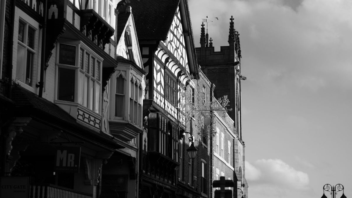 A black and white photograph showcasing historical timber-framed buildings and a prominent stone church tower under a cloudy sky