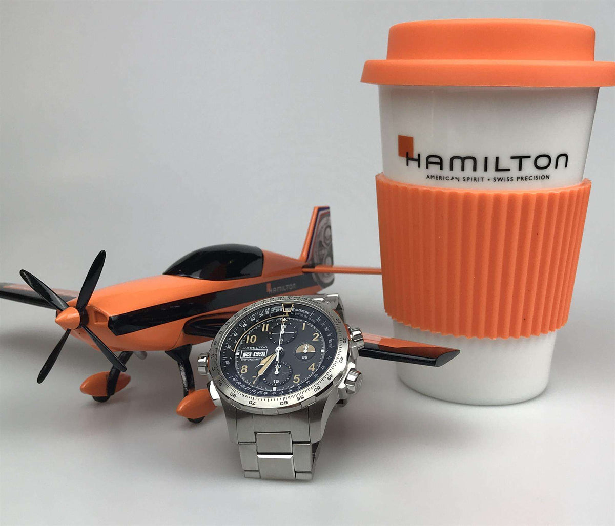 Men's steel chronograph watch with black dial and bracelet beside an orange airplane and tumbler