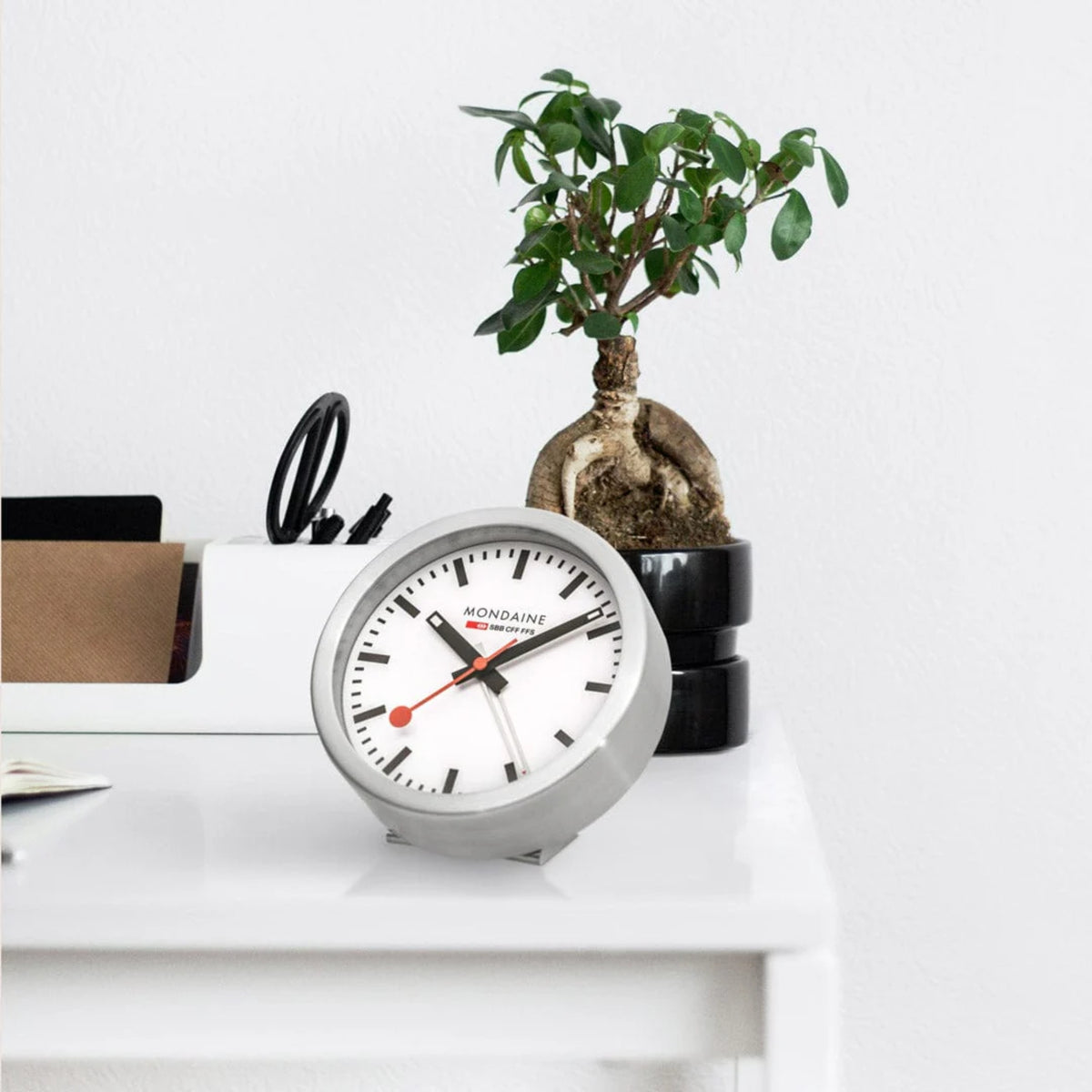 Mondaine Official Swiss Railways Alarm Clock A997.MCAL.16SBB.1 sitting on a white desk next to a potted plant