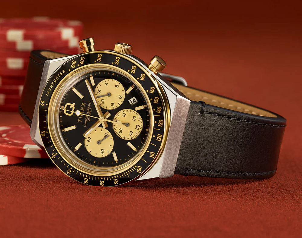 Q Timex chronograph watch with black dial, gold sub-dials, tachymeter bezel, and black stitched leather strap