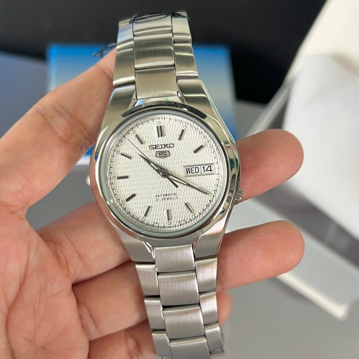 Seiko 5 SNK601K1 mens wristwatch held in hand, displaying a white patterned dial and a steel link strap