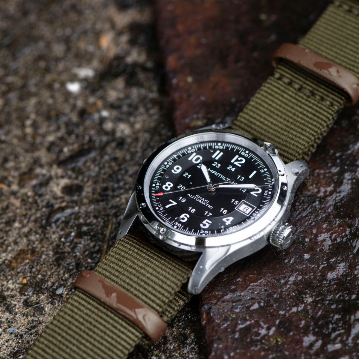 Automatic watch with black dial and olive-green NATO strap on textured surface