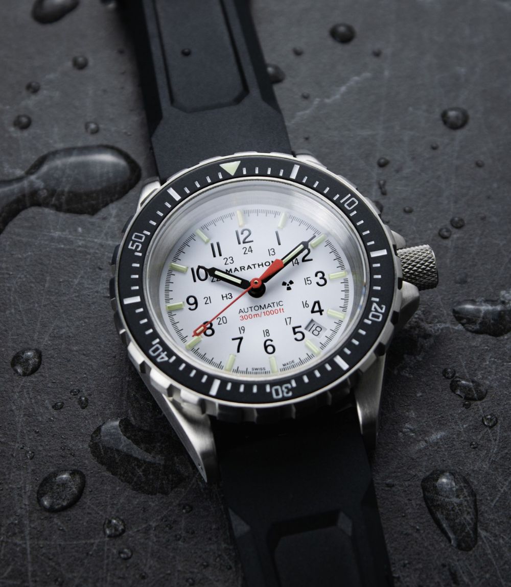 A close-up of a Marathon watch with a silver stainless steel case, white dial, black bezel, and black rubber strap, resting on a dark wet stone surface