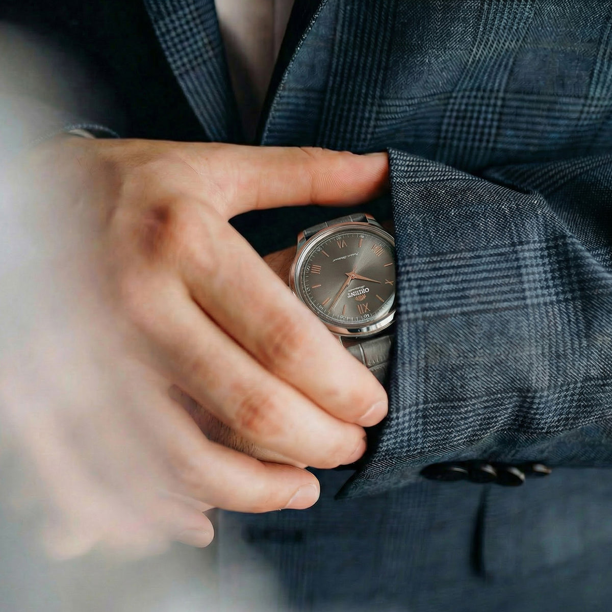 Close-up of a man wearing a grey Orient dress watch styled under a navy plaid suit jacket sleeve