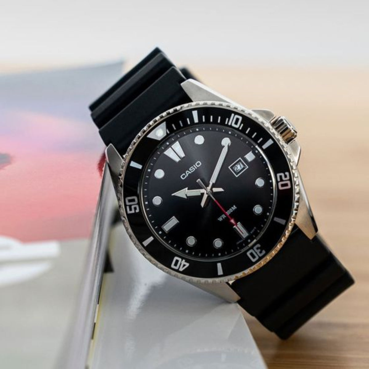 Casio Timeless MDV-107-1A1VEF resting against a magazine on a wooden surface next to a notebook