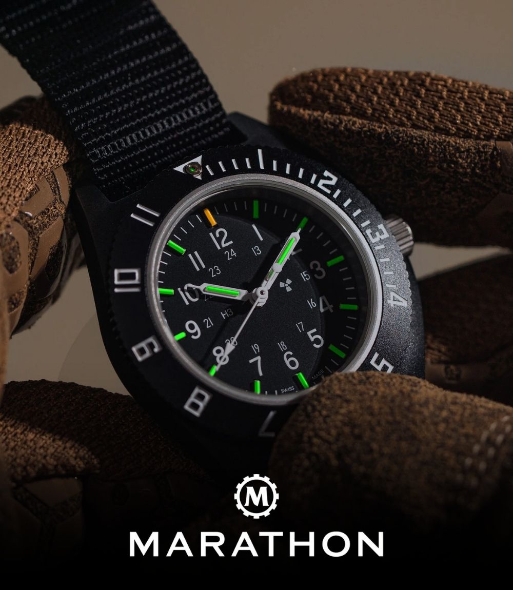 Marathon tactical military watch held in gloved hands, featuring a black case, black dial with luminous green markers, a rotating bezel, and a black nylon strap, featuring the Marathon logo at the bottom