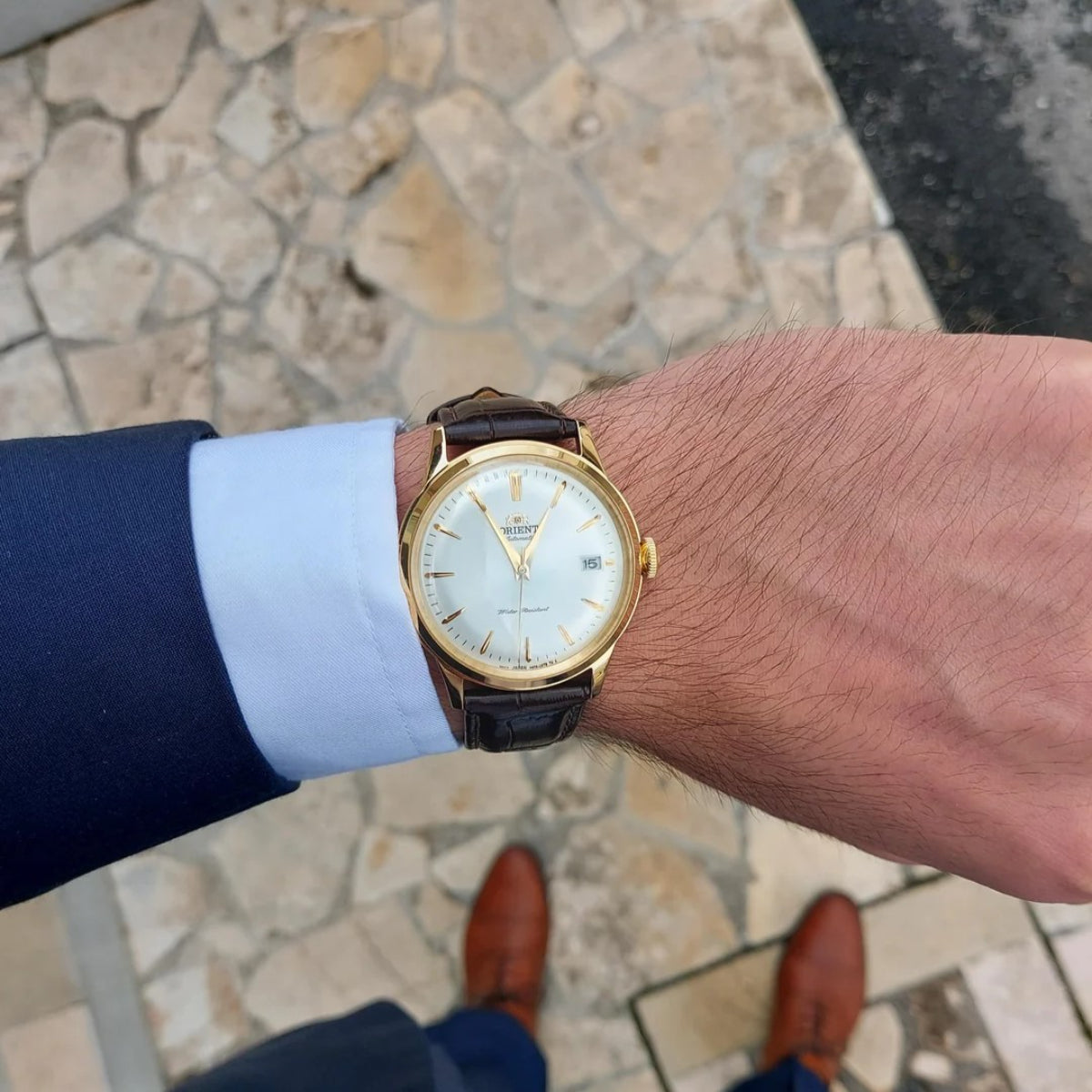 Orient Bambino RA-AC0M01S30B gold-tone wristwatch on a person's wrist with a stone pavement background