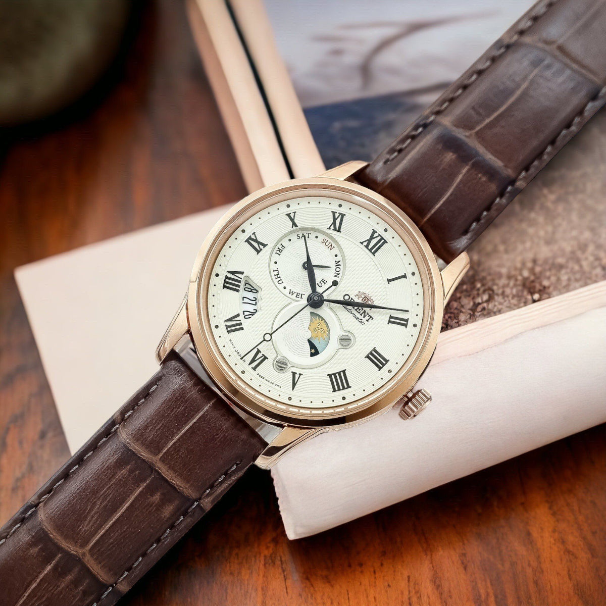 Orient Bambino Sun & Moon RA-AK0007S30B mens wristwatch displaying a champagne dial and rose gold case lying across a wooden surface