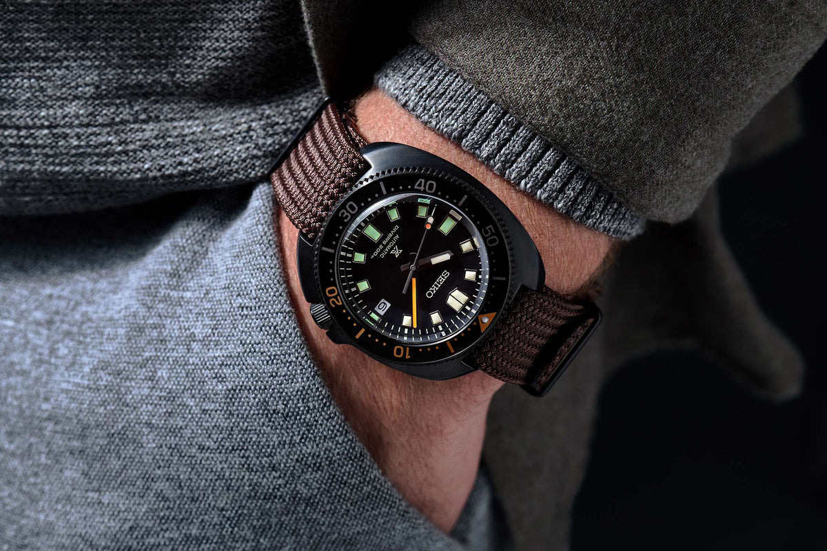Wrist-worn black dial dive watch with orange bezel accents and brown textile strap