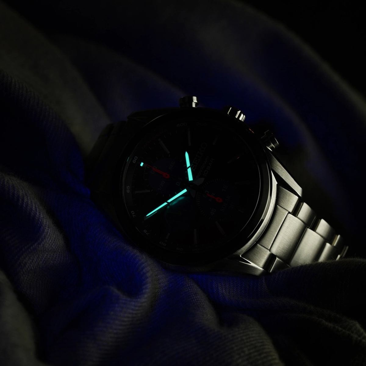 Solar chronograph wristwatch with black dial, luminous turquoise hands, steel bracelet