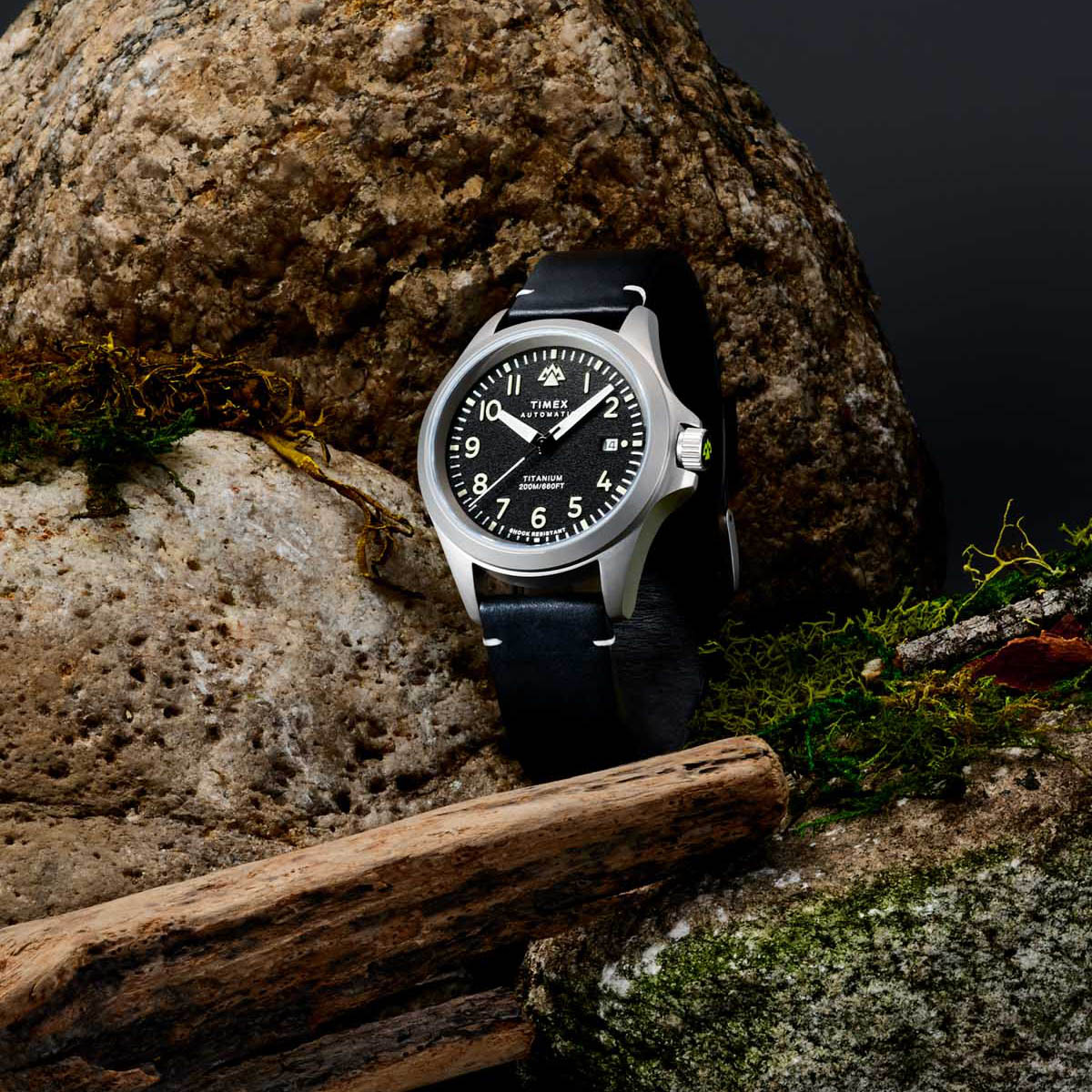 Timex Expedition titanium automatic field watch with black dial and leather strap set against mossy outdoor rocks