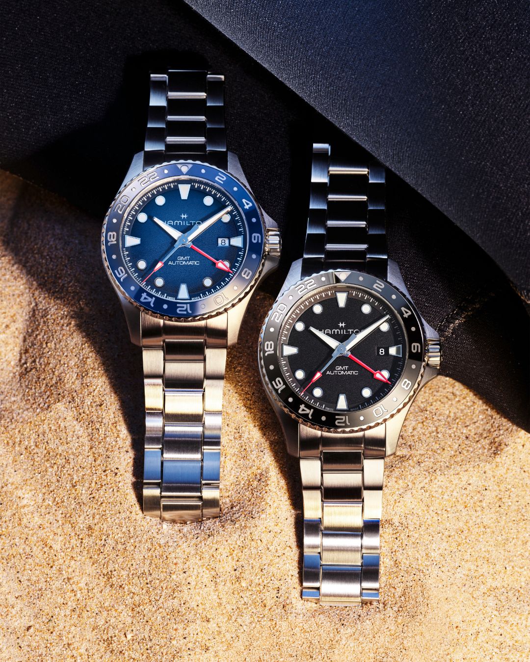 Two Hamilton Khaki Navy Scuba GMT automatic dive watches with blue and black dials displayed on sand