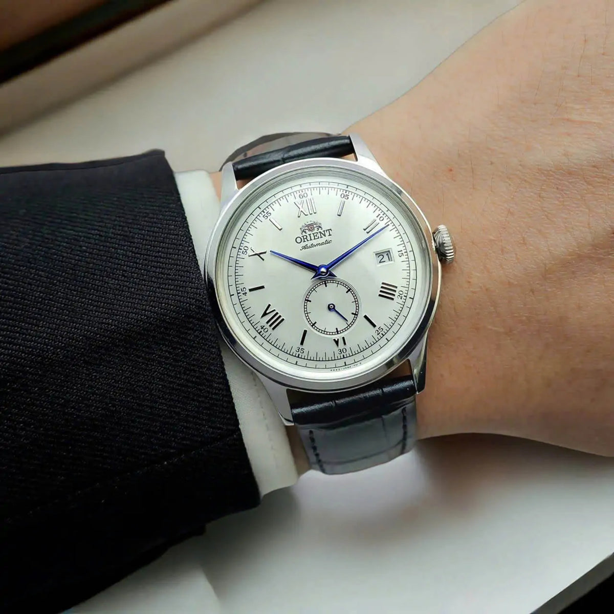 Wrist shot of the Orient Bambino RA-AP0104S30B mens dress timepiece with white dial worn with a black suit jacket