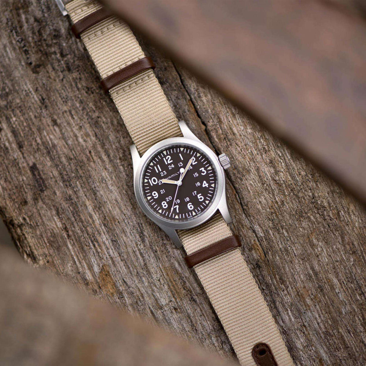 Men’s mechanical field watch on wood surface, dark brown dial, tan NATO strap, brown leather ends.