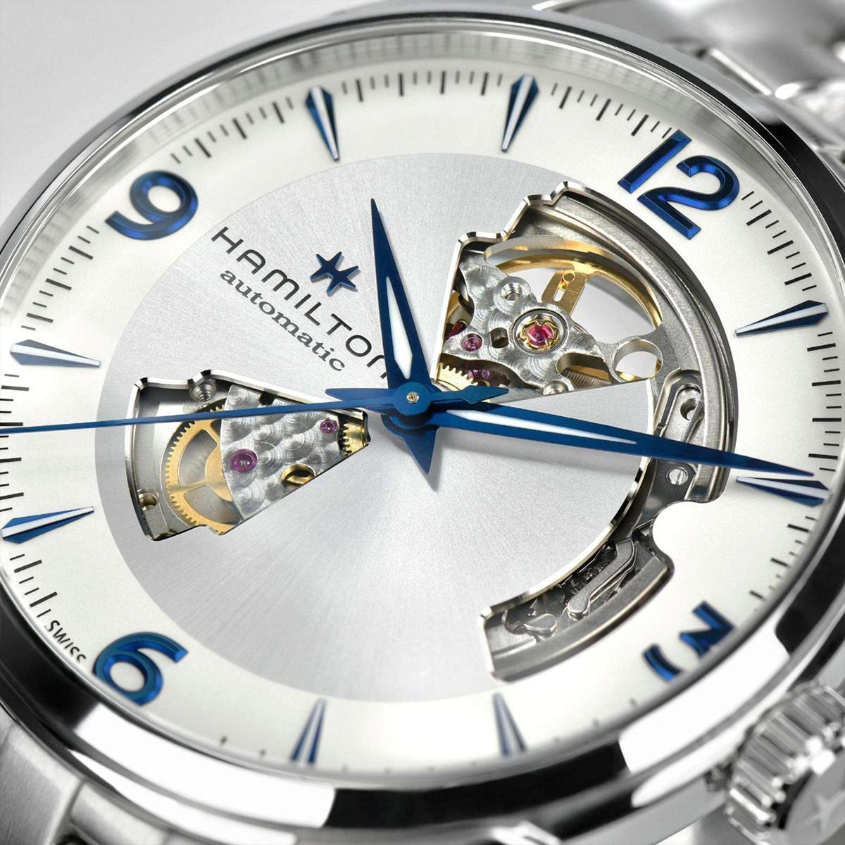 Open-heart watch face close-up with white dial, blue hands, visible gears, silver case