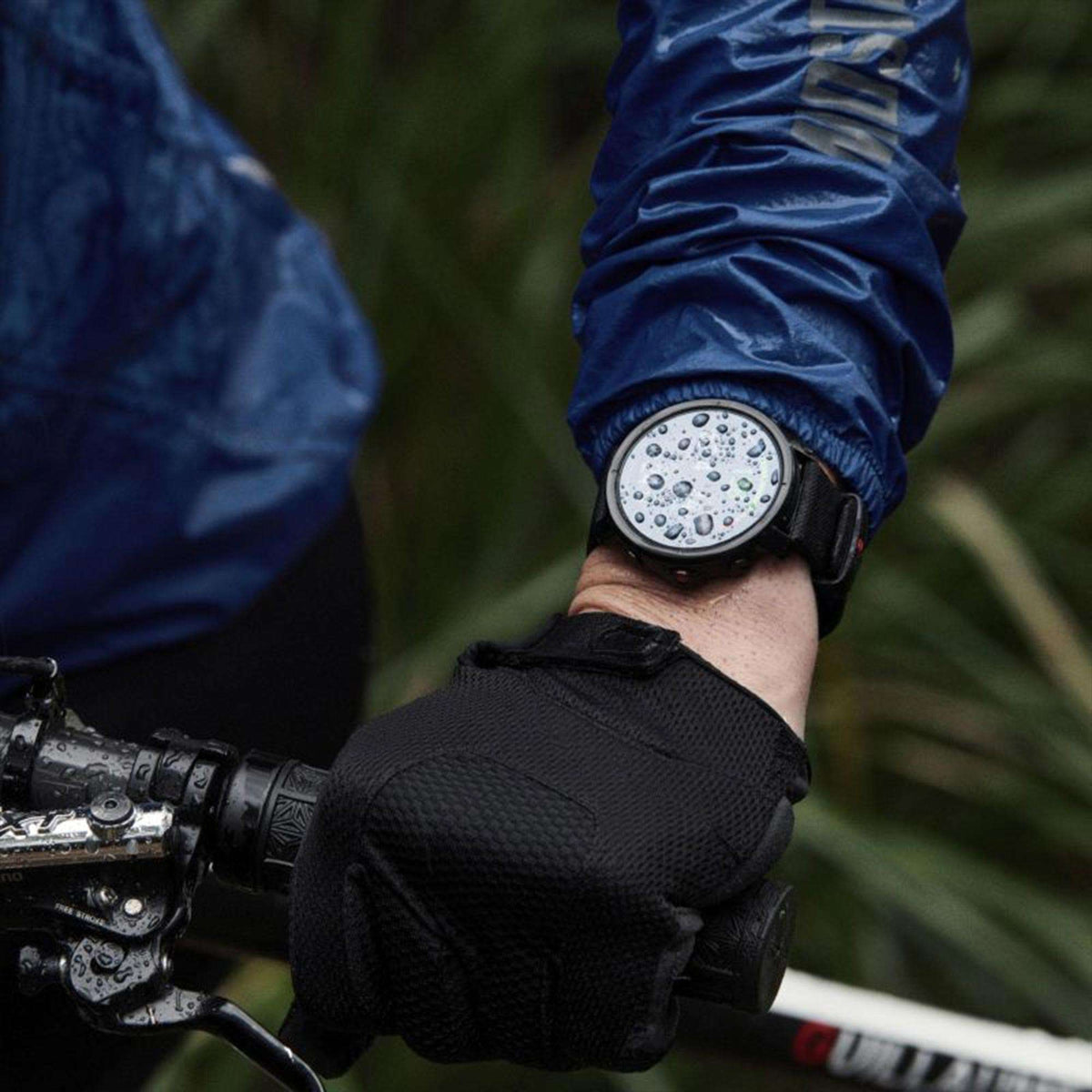 Black smartwatch with rubber strap on wrist, water droplets on face, outdoors background.