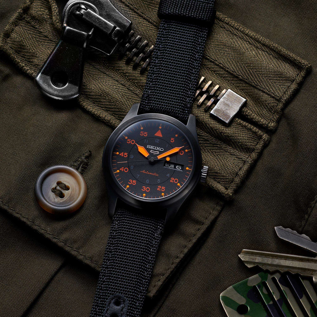 Men's automatic pilot watch with black-orange dial on black strap, angled on olive fabric