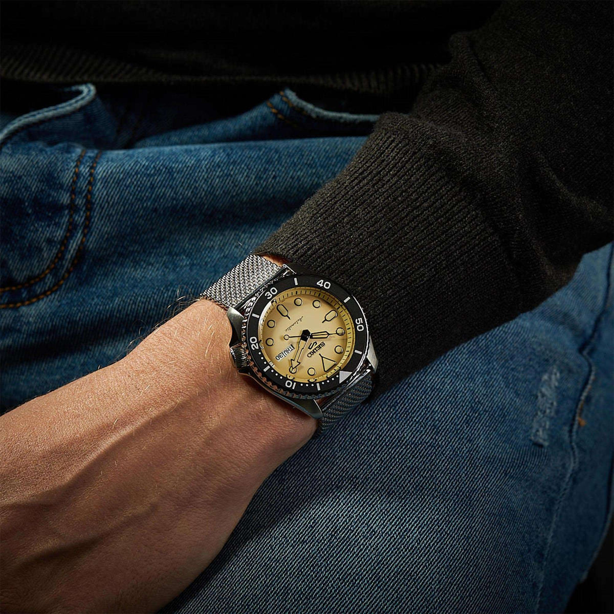 Men's watch on wrist with cream dial, black bezel, and silver Milanese mesh bracelet