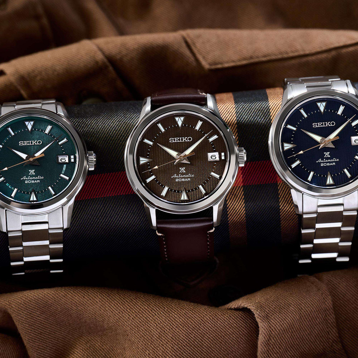 Three watches on fabric; center shows brown dial men's automatic watch with brown leather strap.