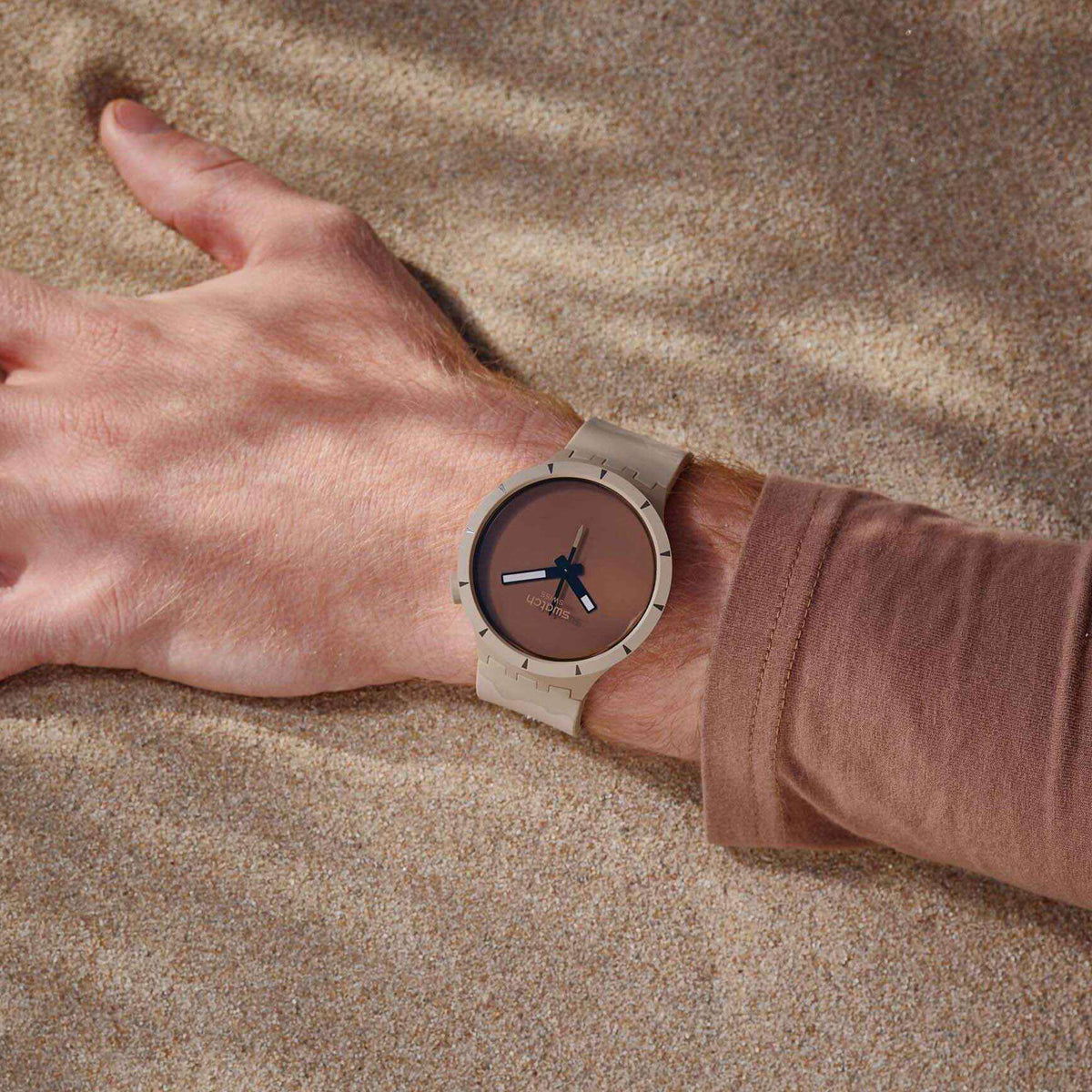 Swatch watch on wrist, beige silicone strap and brown dial with bold black hour markers.