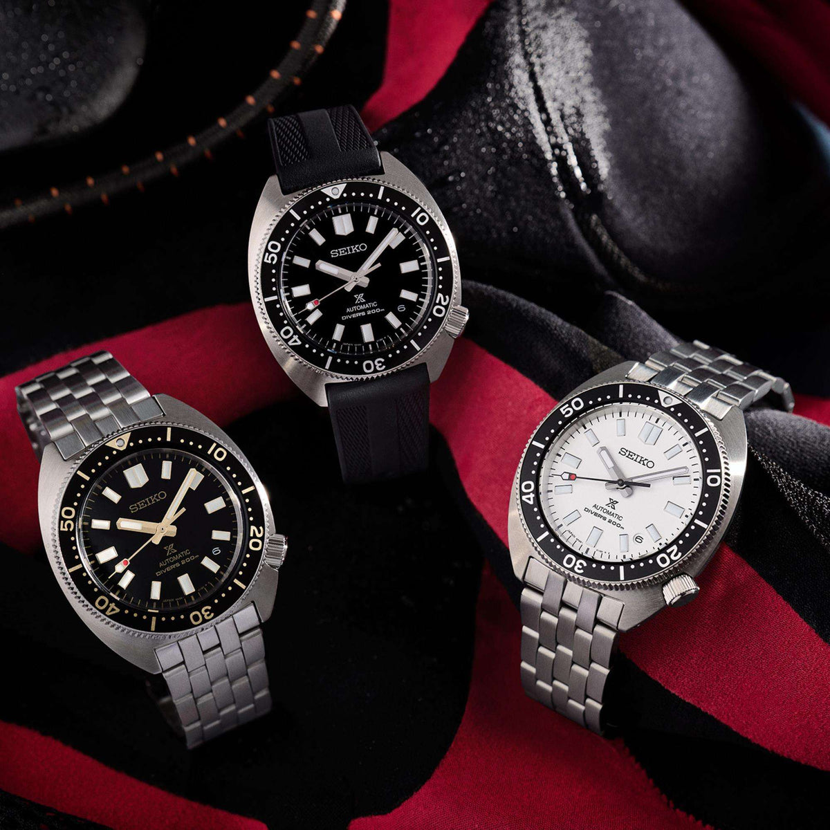 Three Seiko diver watches on red fabric: one white dial, two dark-dial steel bracelets