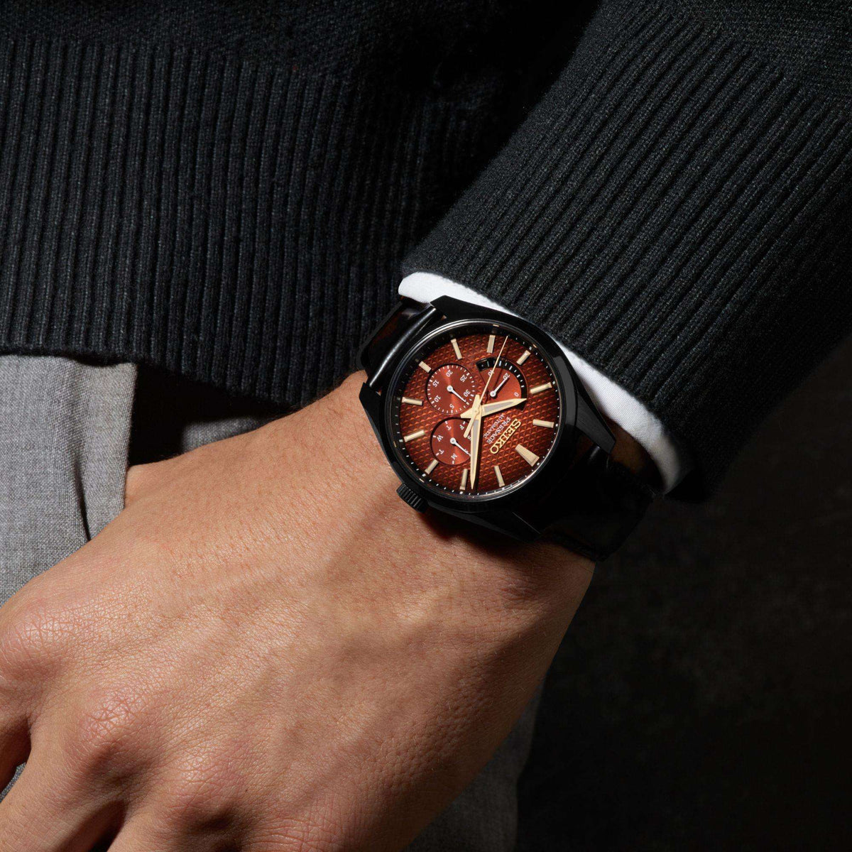 Men's wristwatch: black leather strap on wrist, red-brown dial with gold hands and subdial
