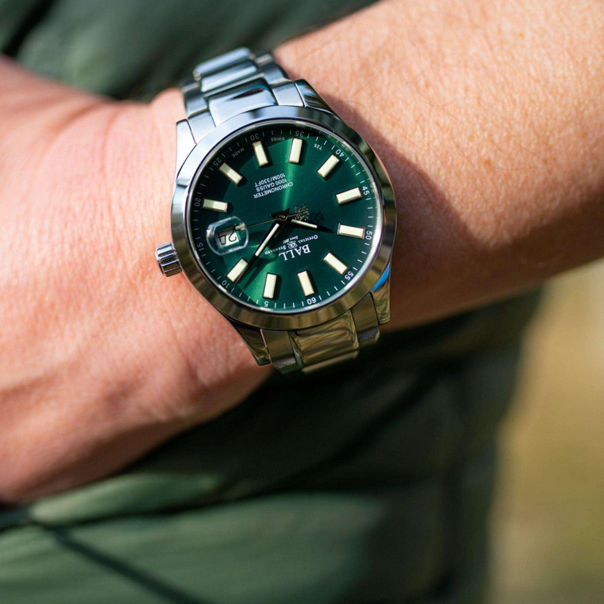 Men's wrist watch on wrist, dark green face with stainless steel bracelet and date window.