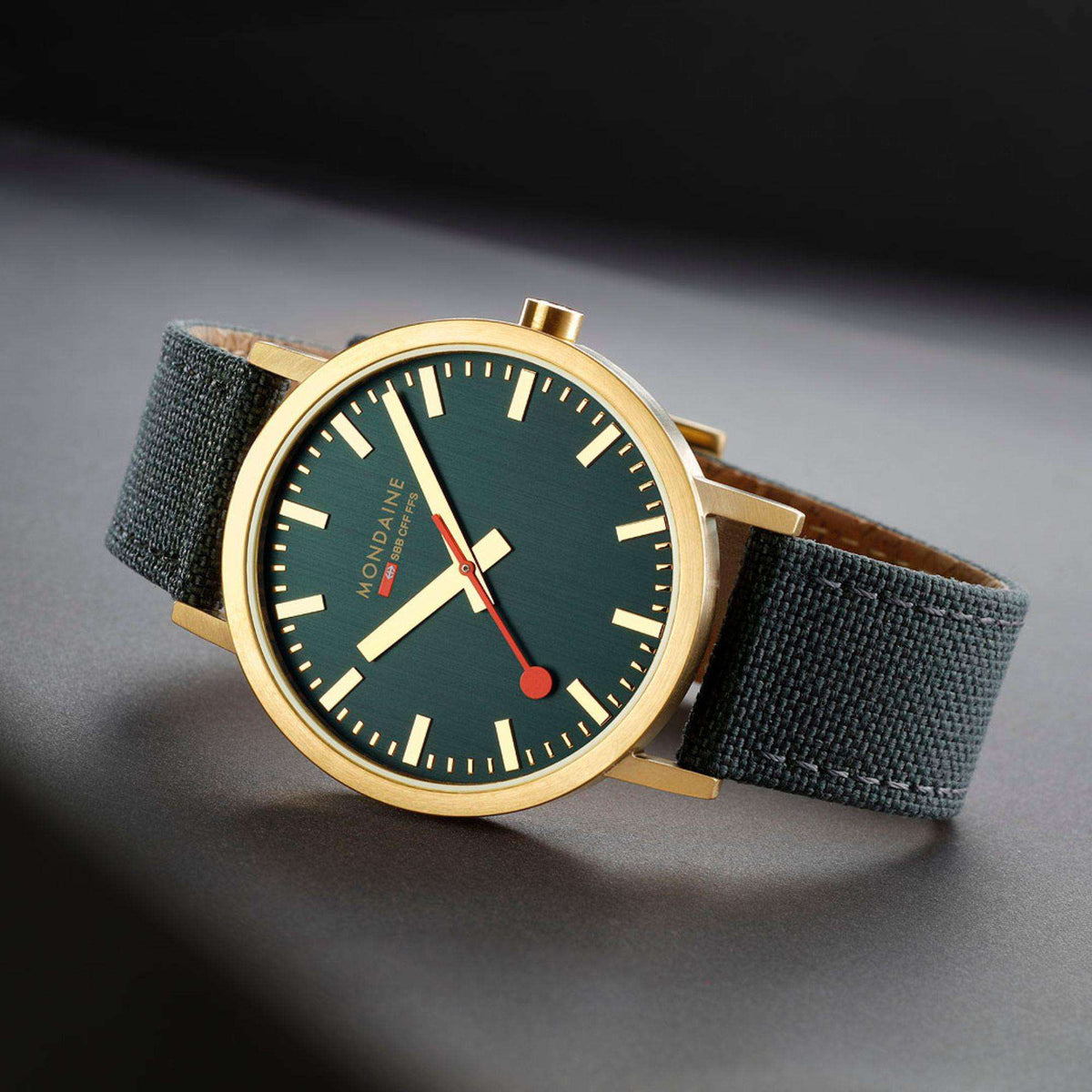 Close-up of gold-tone watch with dark green dial, red accents, and green fabric strap
