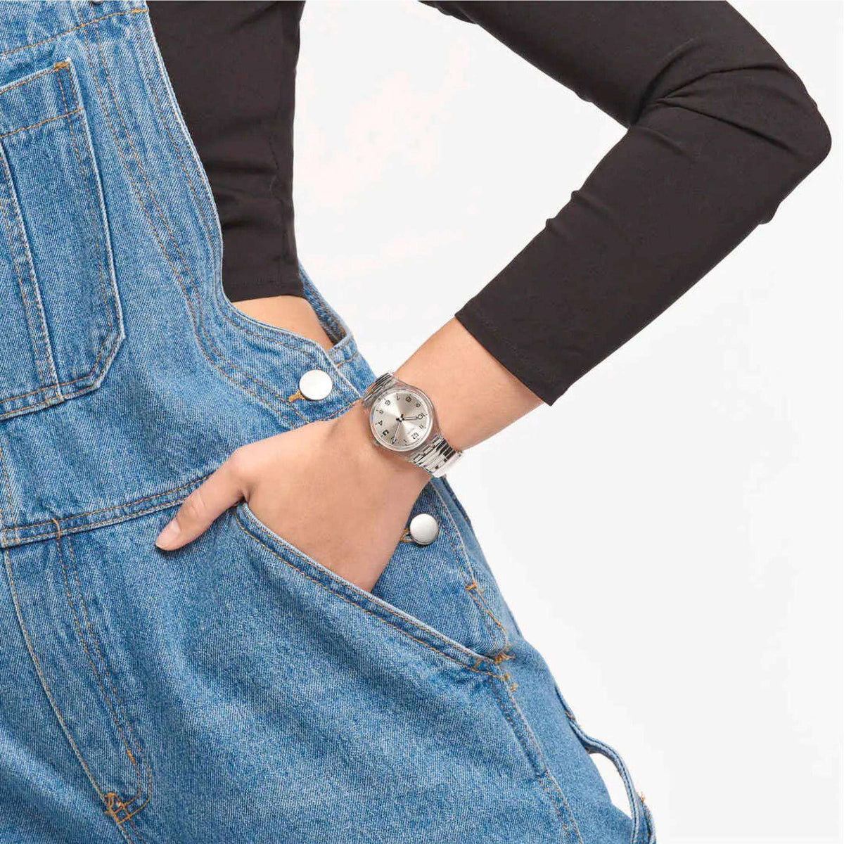 Quartz watch on wrist with silver metal bracelet and round analog face over denim overalls