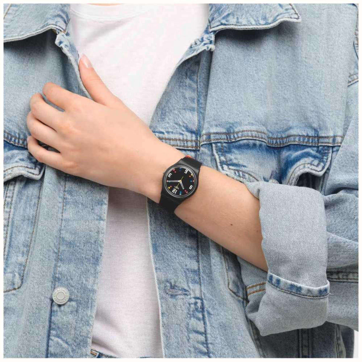 Swatch wristwatch on wrist with black strap and rainbow stripe, multicolor dial against denim.
