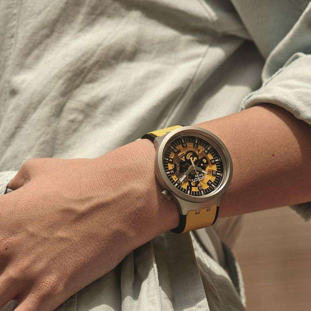 Men's Swatch wristwatch with yellow strap, black dial, and gray case worn on wrist.