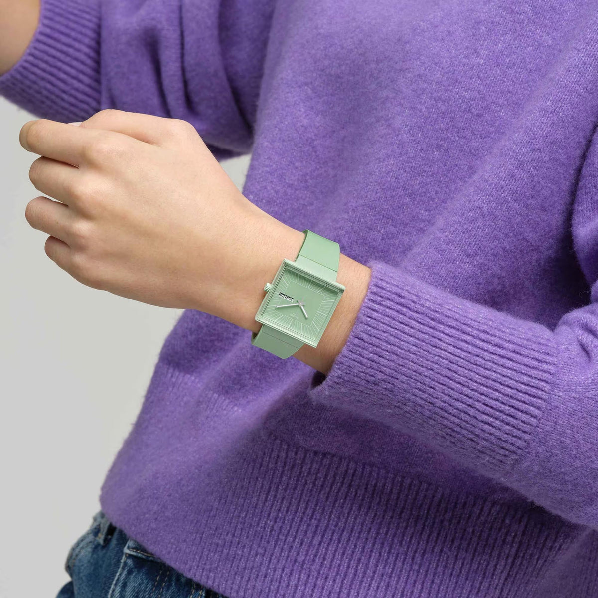 Mint green square wristwatch on wrist over a purple sweater sleeve