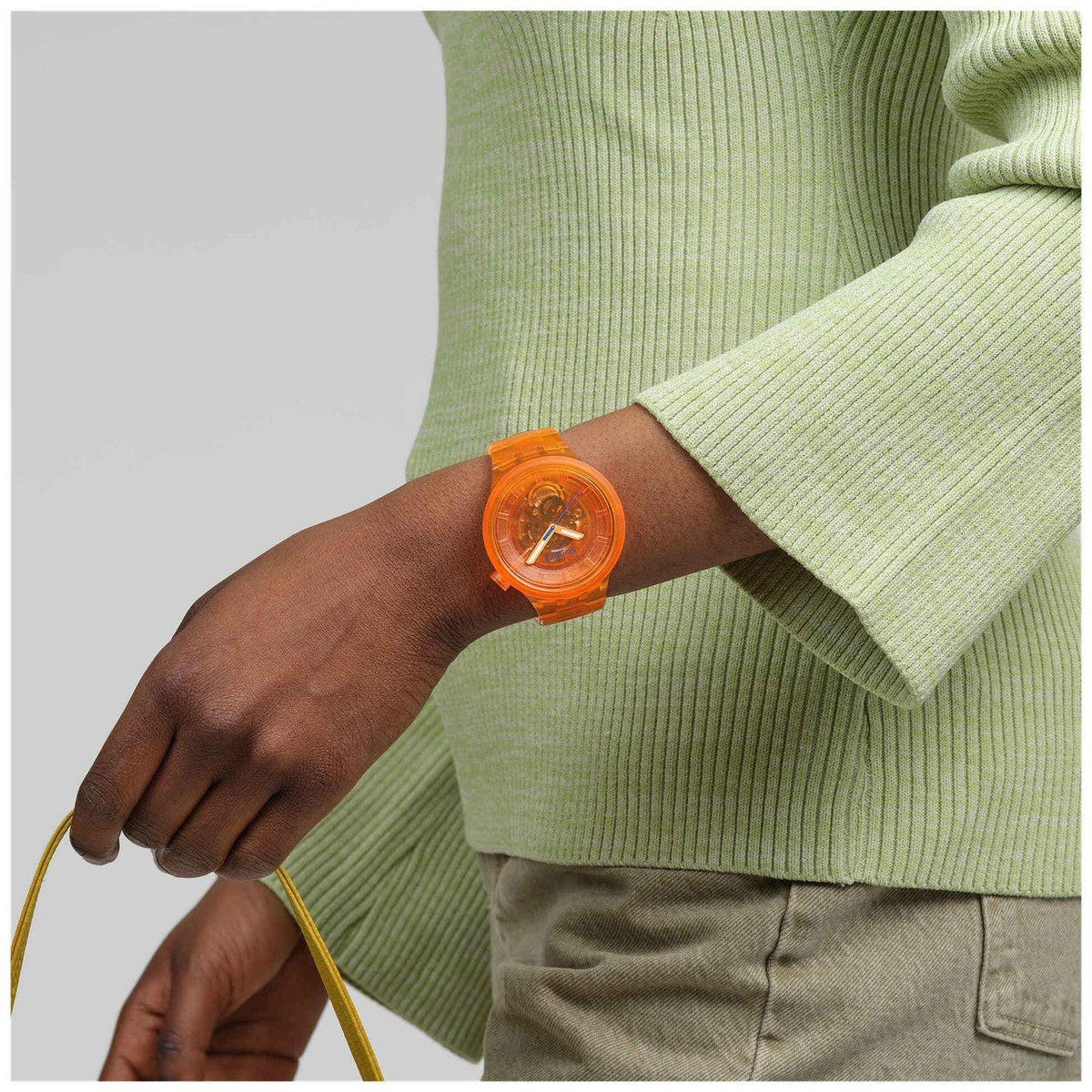 Orange Swatch bio-sourced watch on wrist with orange dial, worn with green knit sweater.