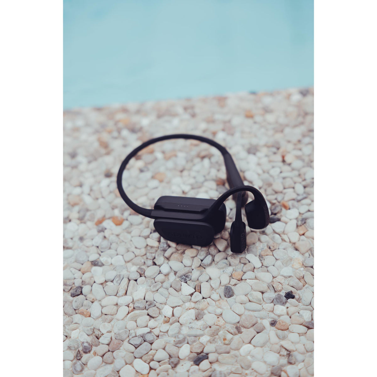 Suunto Aqua SS051023000 black sports open-ear wireless headphones resting on pebble surface showcasing portable charging case