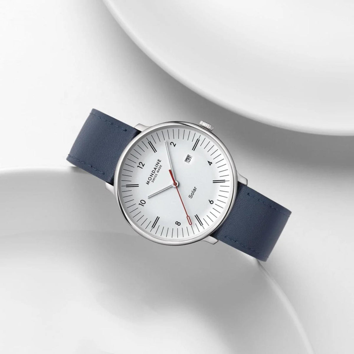 Mondaine watch with silver case and blue leather strap angled between two curved white ceramic plates