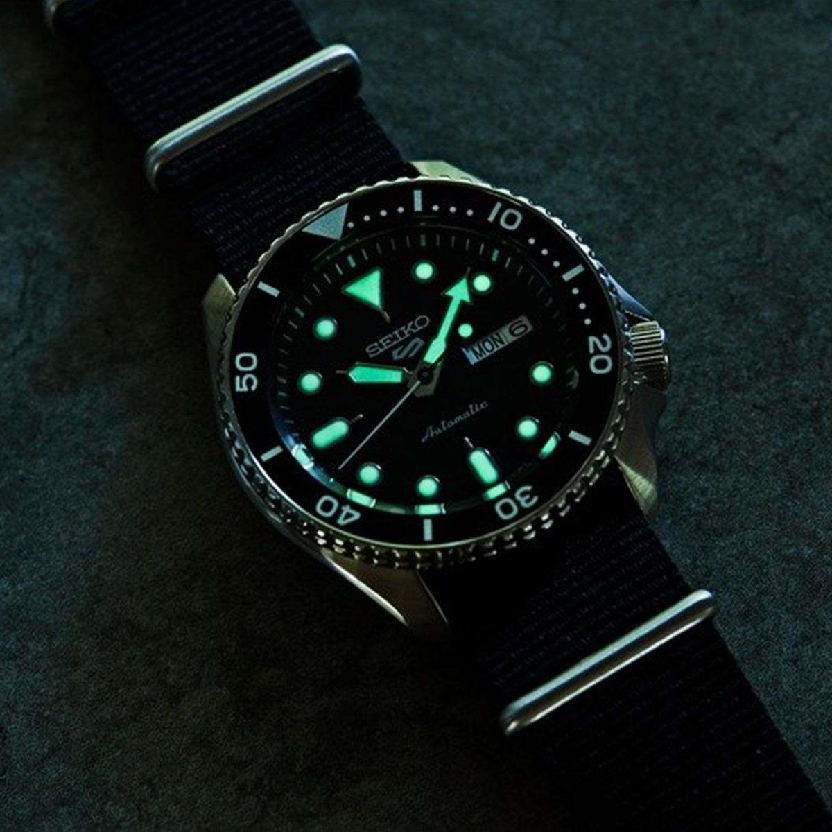 Men's automatic watch with glowing green markers on black dial and black fabric strap.