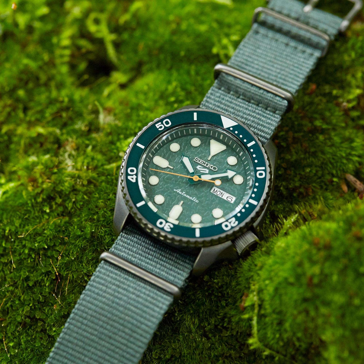 Men's automatic watch with green dial, stainless case, and light green fabric strap.