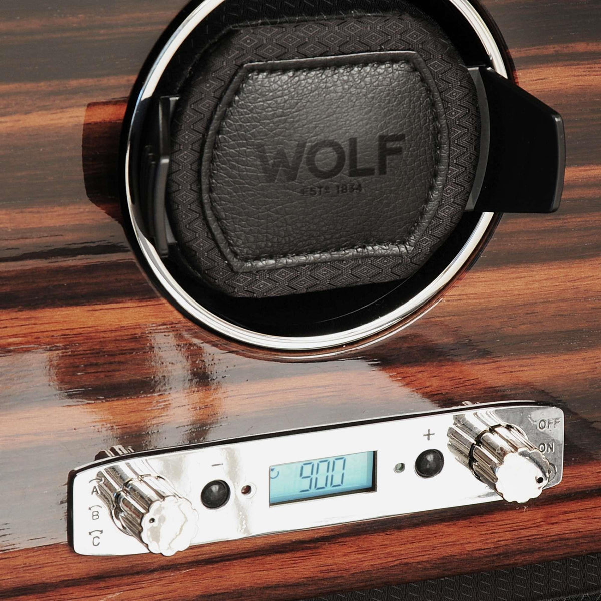 Close-up of watch winder cushion with brand embossing and silver control knobs on wood panel