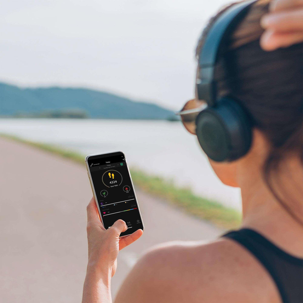 Rose gold smartwatch user checking activity on smartphone near a lakeside path