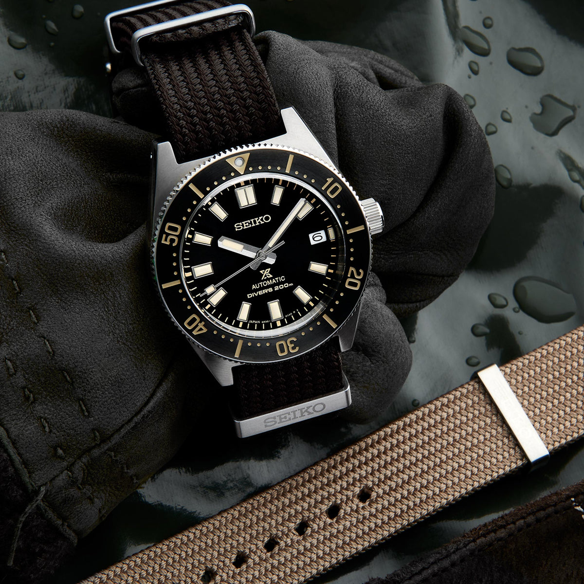 Men's Seiko diver watch on glove beside tan strap, black dial with gold indices