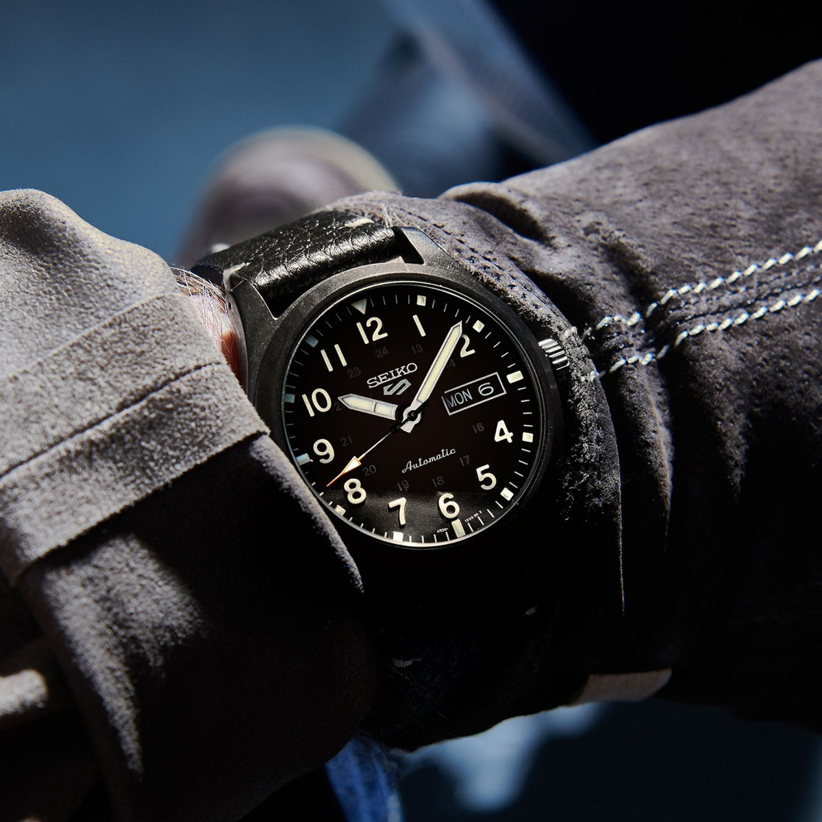 Black field watch worn on wrist, black leather strap, day-date at 6 and bold numeral markers