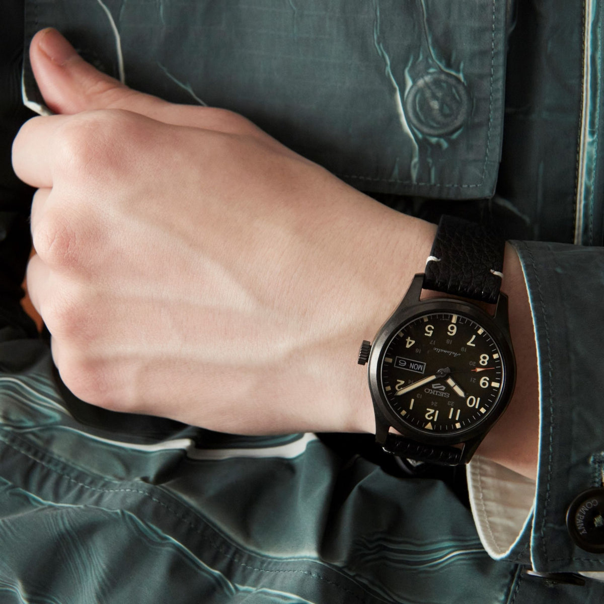 Black field watch on wrist with black leather strap, day-date window, and glowing numeral indices