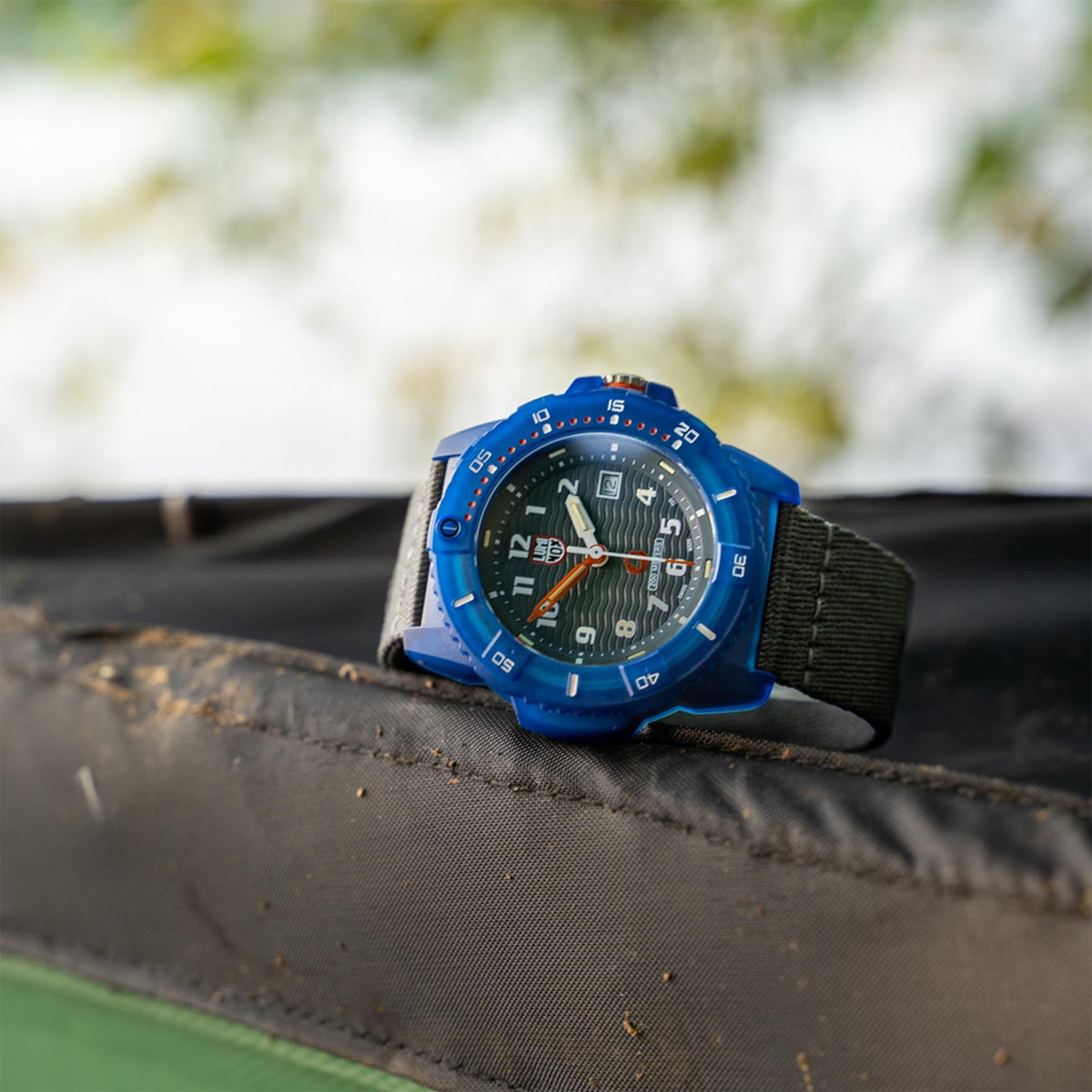 Men's Luminox watch lying on surface: blue case, grey strap, black dial with date window