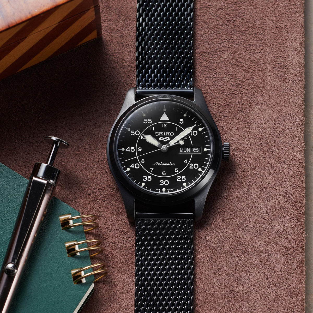 Black Seiko watch with mesh band on a textured surface with pen and notebook.