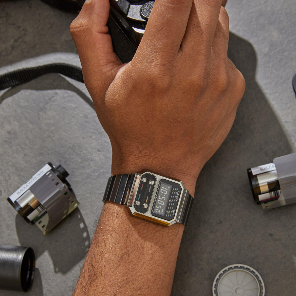 Grey digital men’s watch with square case, metal bracelet, LCD display on wrist.
