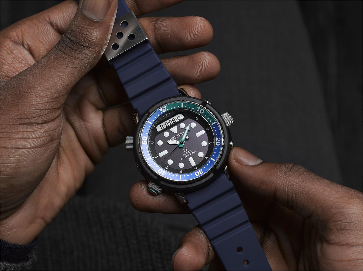 Close-up of watch in hand: blue silicone strap, black dial, blue bezel, green accents.