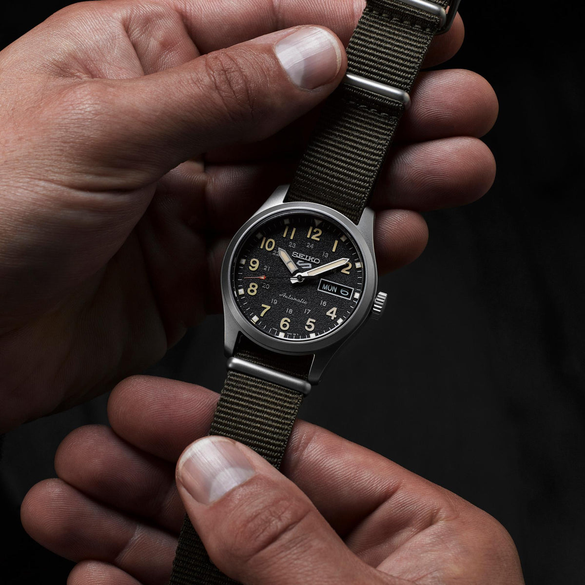 Automatic watch with black dial on olive NATO strap, held in a hand