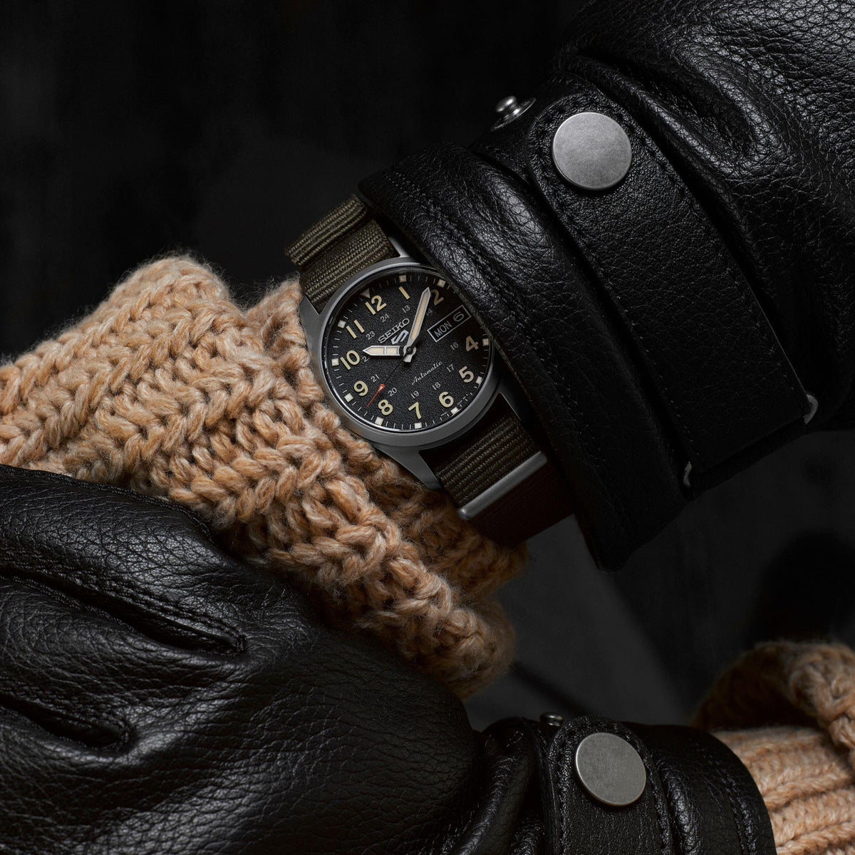 Automatic watch with black dial and olive NATO strap resting on black leather gloves